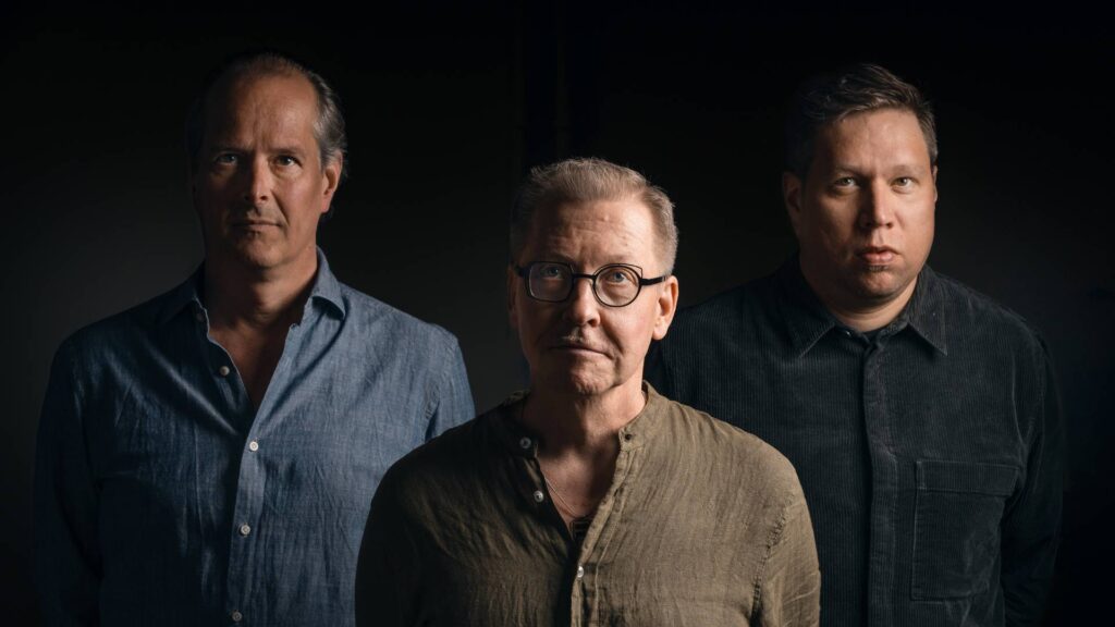 Kolme miestä seisoo kameraan katsoen tummaa taustaa vasten.